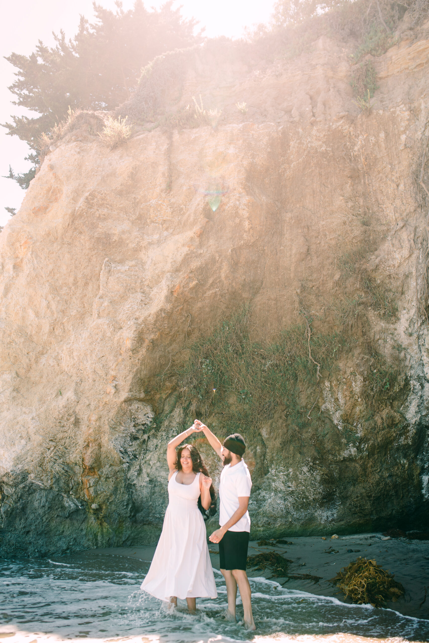 Engaged couple in cream outfits embracing against sandstone cliffs at a Santa Barbara beach engagement session